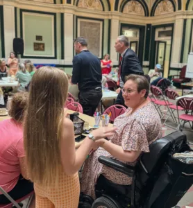 a large hall room with multiple tables inside with many people sat at them talking at an event in the background. In the foreground is a woman in a pink dress sat down in a motorised wheelchair looking to her left and talking to a child with her back to the camera with long blonde hair.