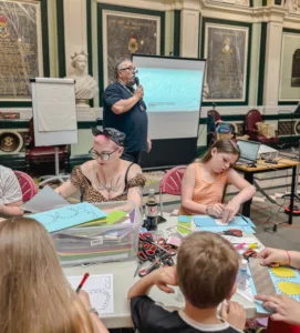 a photograph of a council hall. In the forerground are adults and children sitting around a table doing various crafts busy. In the background Leeds GATE staff member Robin stands looking to the right with a projector screen behind him addressing the room about policy. He wears a Leeds GATE t shirt.