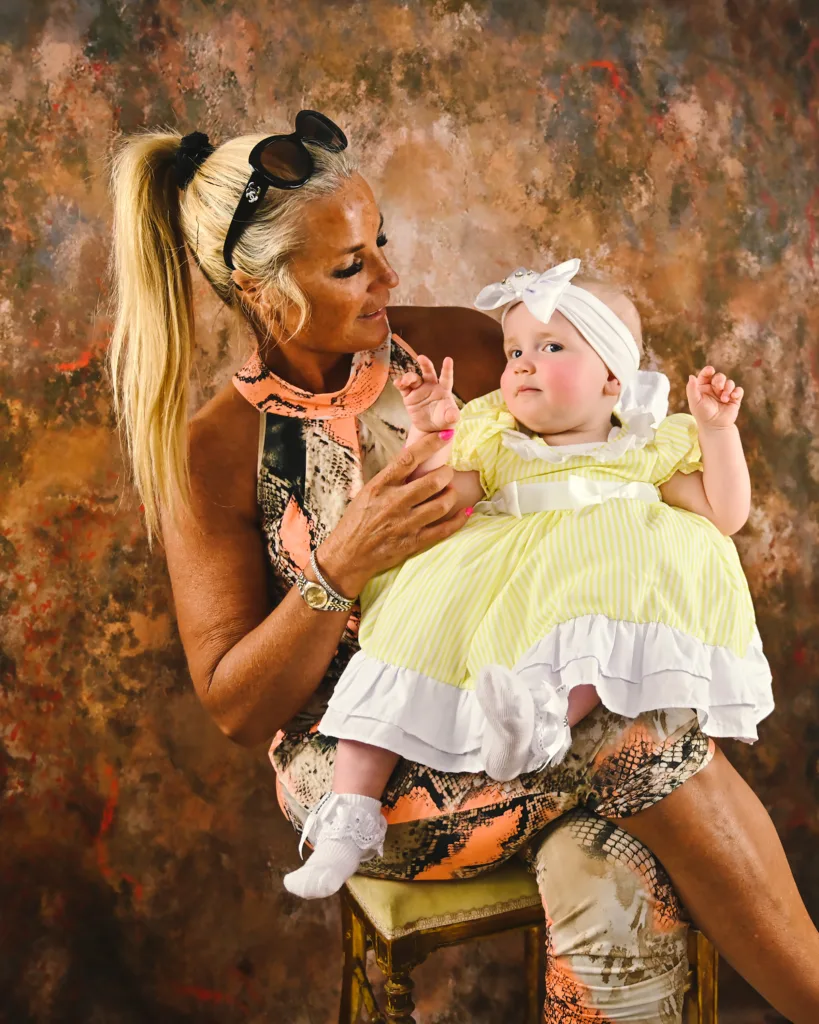 A woman with long blonde hair in a ponytail and dress sits in front of a brown background with a baby in a yellow dress and bow headband on her lap