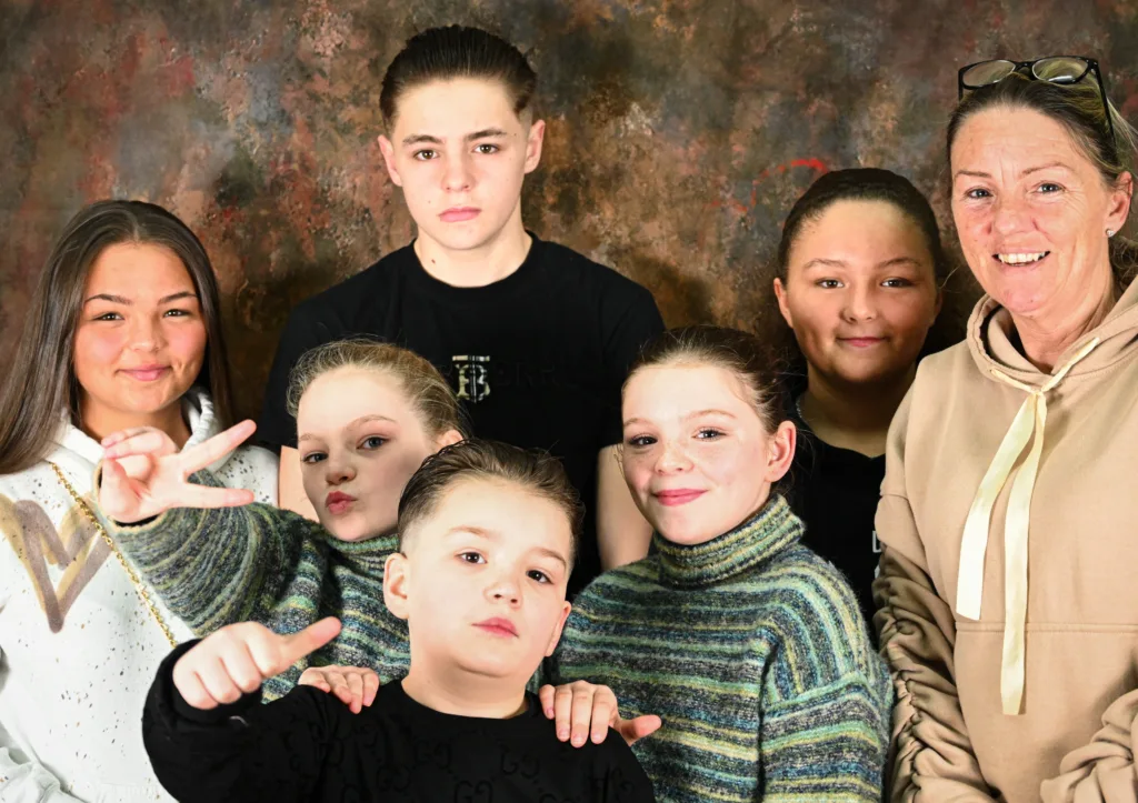 A family stands in front of a photography background with a mother smiling on the right with six children smiling and doing poses like thumbs up and peace signs