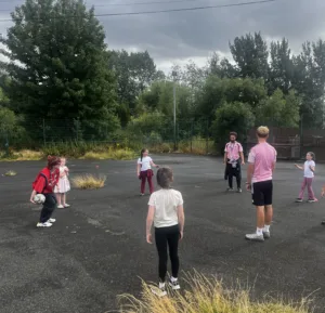 Leeds United foundation coaches on a flat tarmac surface in a circle with young people and a youth worker throwing a football for them on a Gypsy and Traveller site